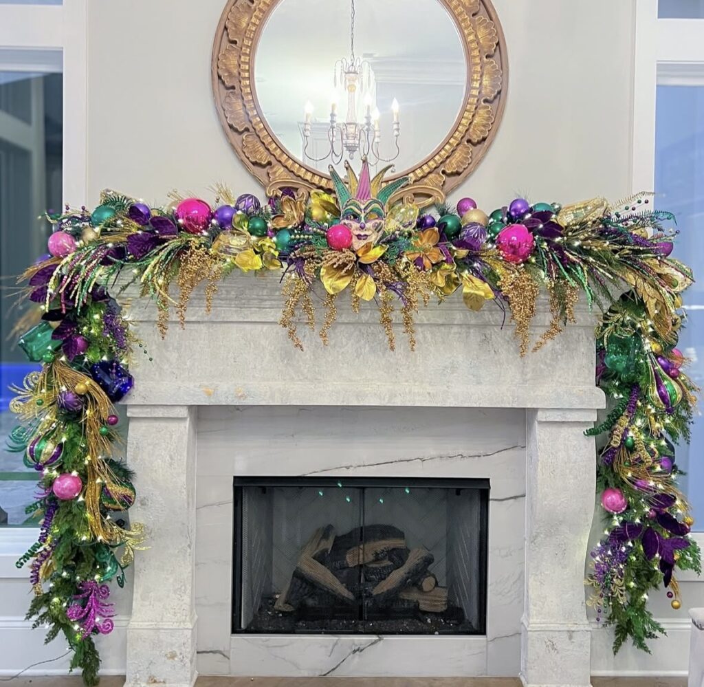 Stone fireplace mantel dressed in Mardi Gras colors with a full garland of purple, green, and gold ornaments, a jester face centerpiece, twinkling lights, and cascading greenery on both sides.