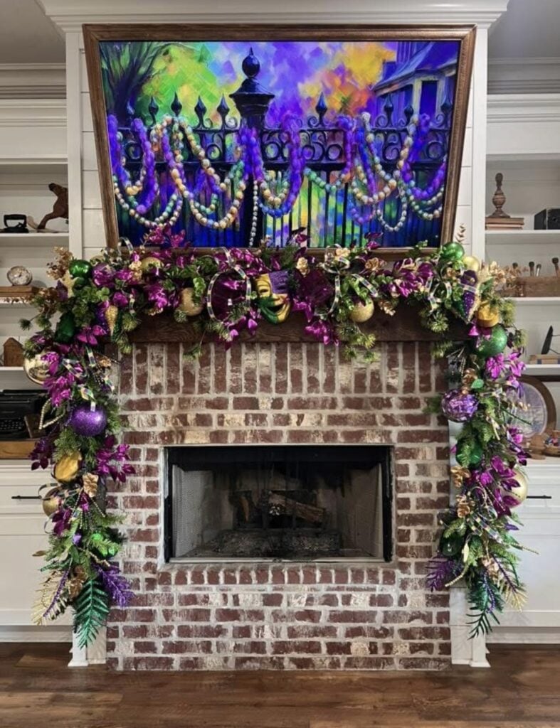 White fireplace mantel with a Mardi Gras garland, gold fringe, purple-green-gold ornaments, fleur-de-lis accents, and big striped bows on both ends.