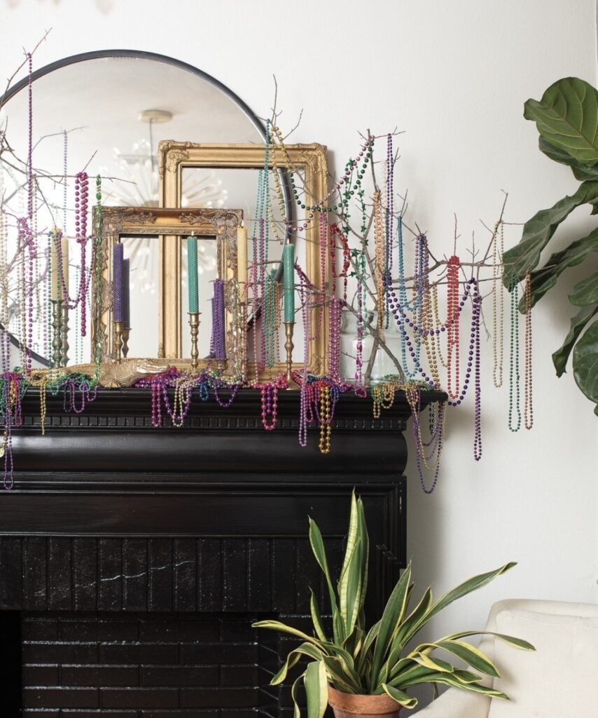 Black mantel Mardi Gras display featuring layered gold frames, colorful bead necklaces draped across branches, and tall taper candles in jewel tones.