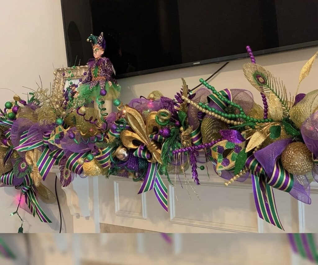 Close-up Mardi Gras mantel garland with purple mesh ribbon, gold and green accents, bead strands, and a small jester doll sitting on the left side near the TV.