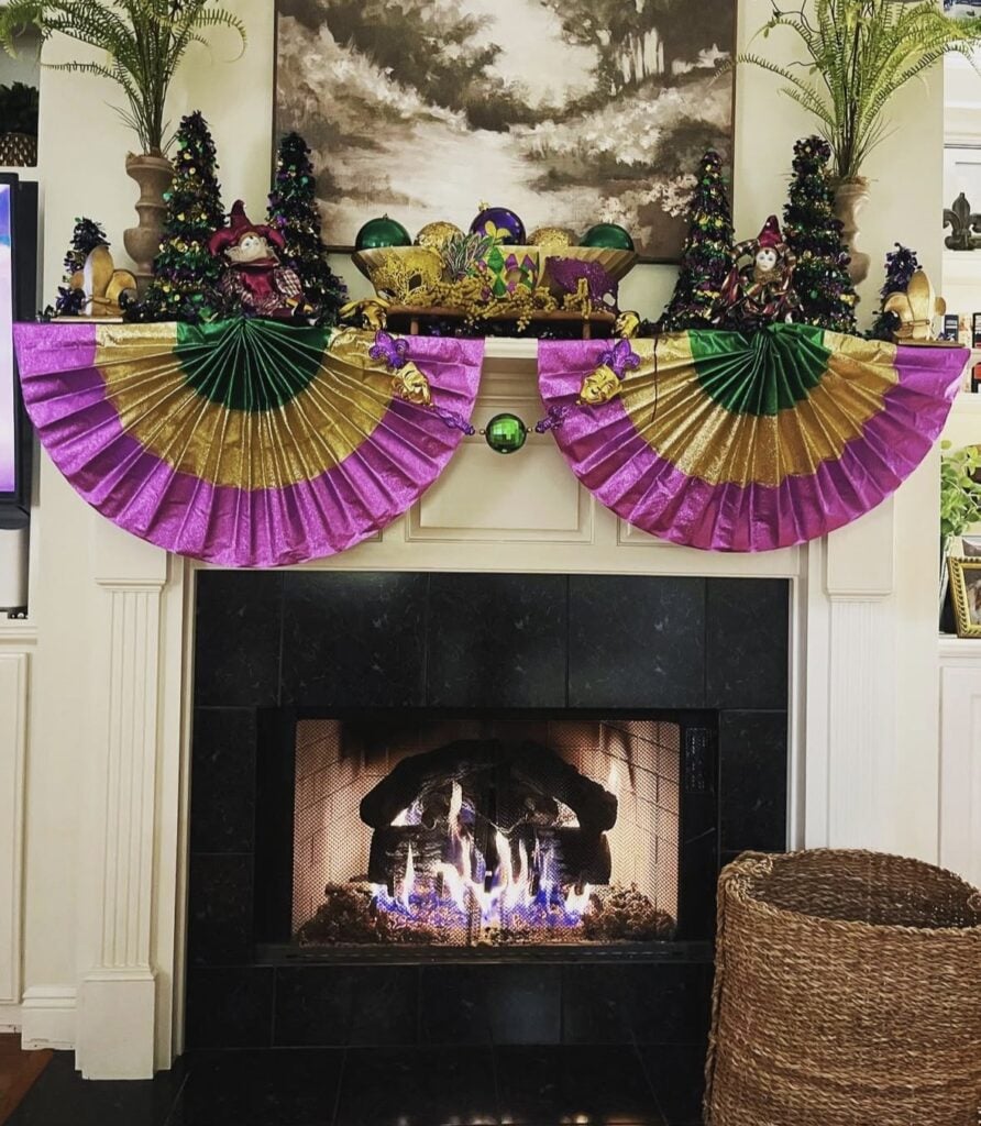 Fireplace mantel with Mardi Gras bunting fans in purple, green, and gold, plus small cone trees, jester figures, and festive table decor arranged across the top.