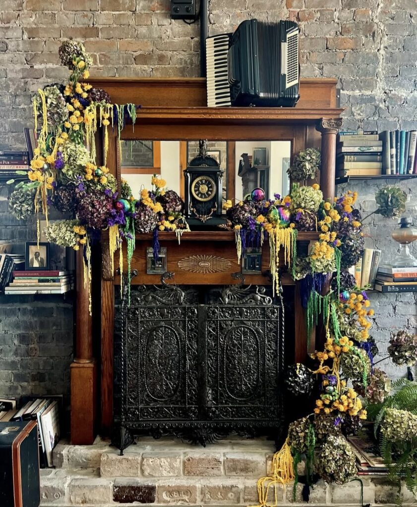 Antique wood fireplace mantel with ornate black fire screen and an overflowing Mardi Gras floral garland on a brick wall.