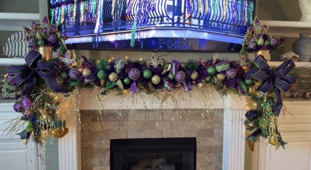 White fireplace mantel with a Mardi Gras garland, gold fringe, purple-green-gold ornaments, fleur-de-lis accents, and big striped bows on both ends.