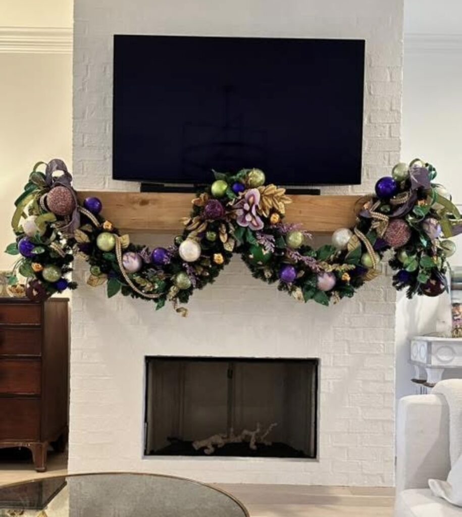 White brick fireplace with a wood mantel under a TV, decorated with three Mardi Gras garland swags in purple, green, and gold.