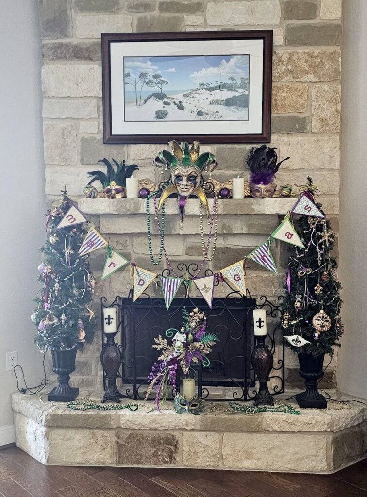 Stone corner fireplace decorated with two small Mardi Gras trees, a jester mask, a “Mardi Gras” banner, beads, candles, and purple-green-gold accents.