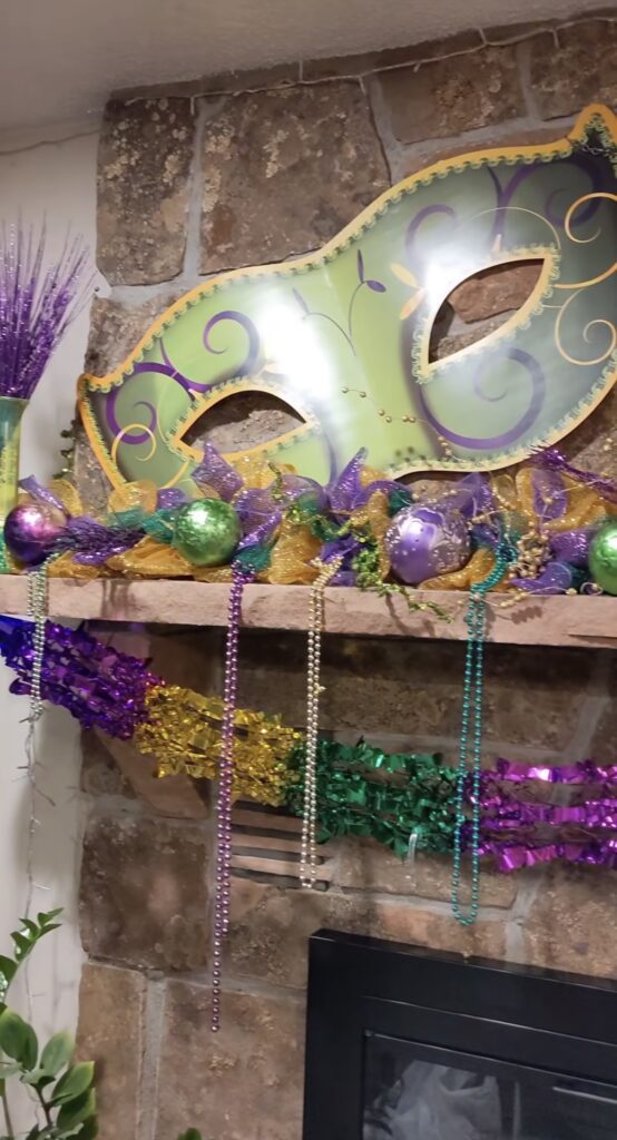 Close-up of a large Mardi Gras mask above a stone fireplace, with purple-green-gold mesh, ornaments, bead strands, and tinsel garland on the mantel.