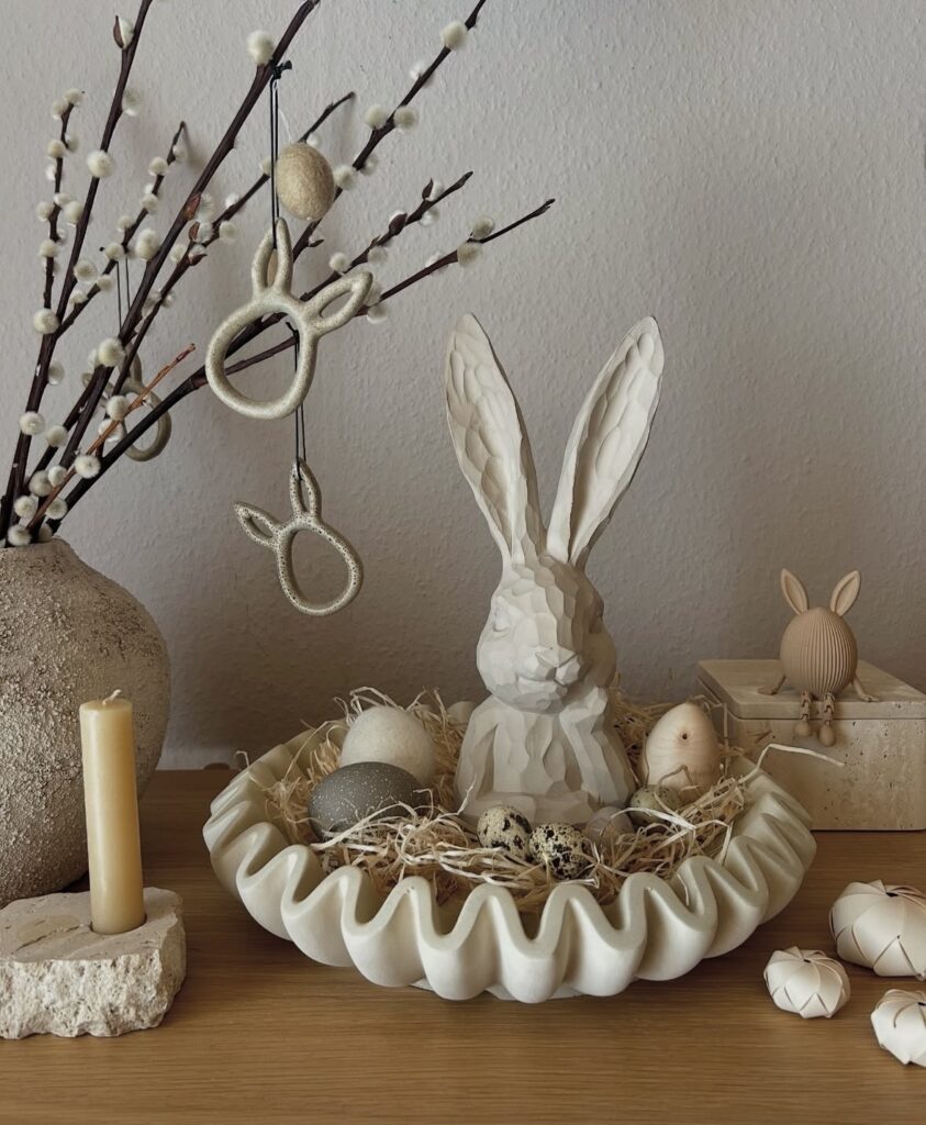 Minimalist Easter decor vignette with a large white bunny statue, decorative eggs in a wavy bowl, and pussy willow branches with hanging bunny ornaments.