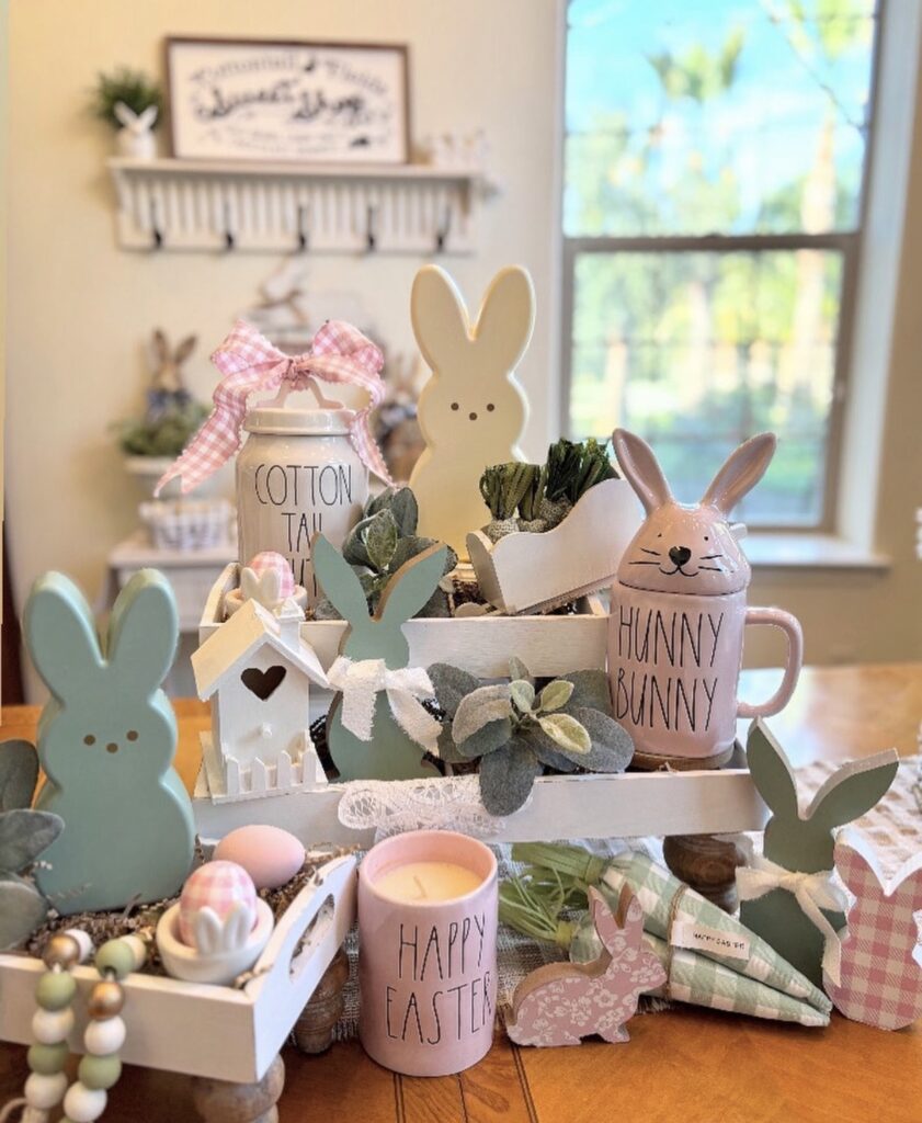 Pastel Easter tiered tray decor with bunny mugs, “Happy Easter” candle, peeps-style bunnies, carrots, greenery, and pink gingham bow.