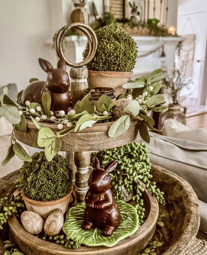 Rustic Easter tiered tray with chocolate bunny figurines, faux greenery garland, topiary balls, decorative eggs, and wooden accents with cabbage leaf plates