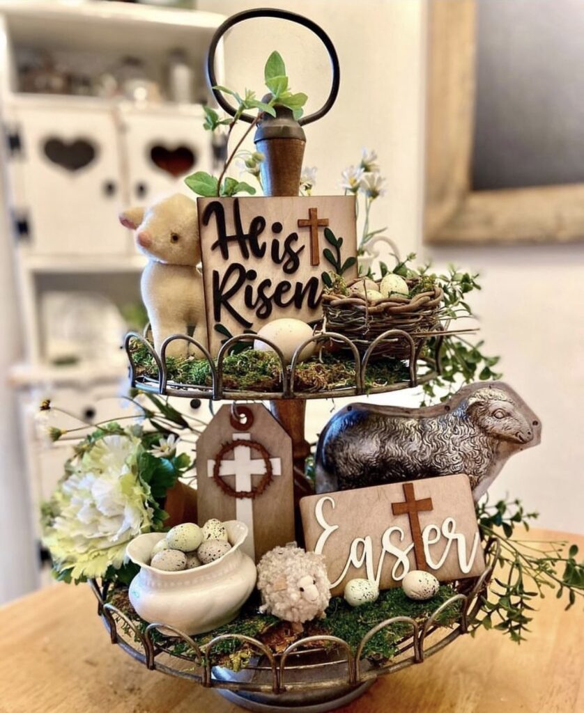 Religious Easter tiered tray decor with “He is Risen” sign, crosses, lamb figurines, moss, speckled eggs, and spring greenery.