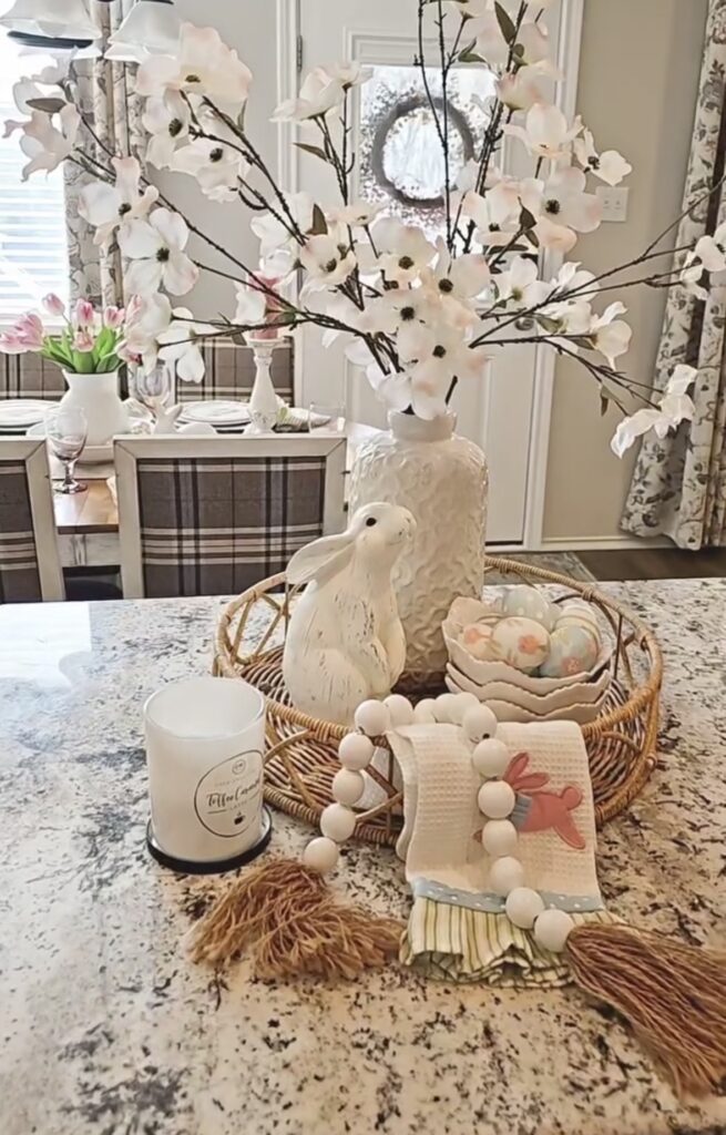 Kitchen island Easter centerpiece tray with magnolia branches in a vase, white bunny decor, stacked bowls of pastel eggs, beaded garland, and a carrot dish towel.