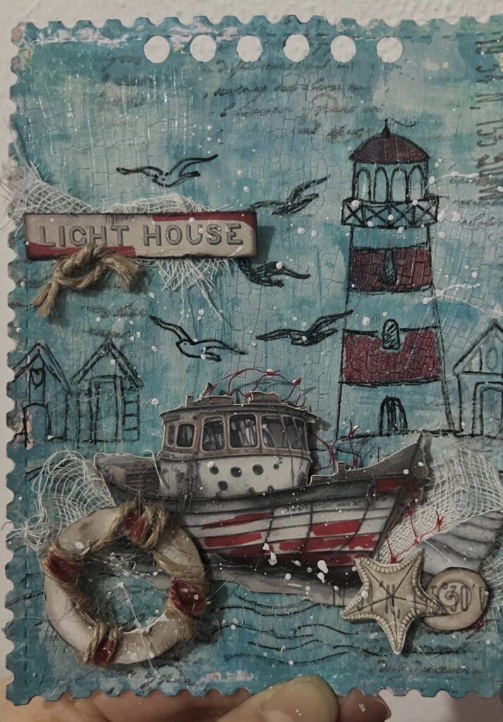 Handmade lighthouse mixed media card with a striped boat, seagulls, netting, and a rope-wrapped life ring on a teal crackle background.
