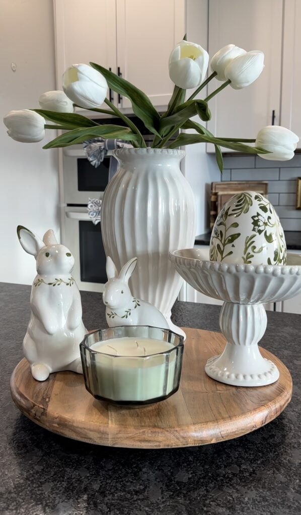 White Easter kitchen decor on a round wooden tray with a white ribbed vase of white tulips, ceramic bunny figurines, a candle, and a white pedestal bowl holding a floral decorative egg.