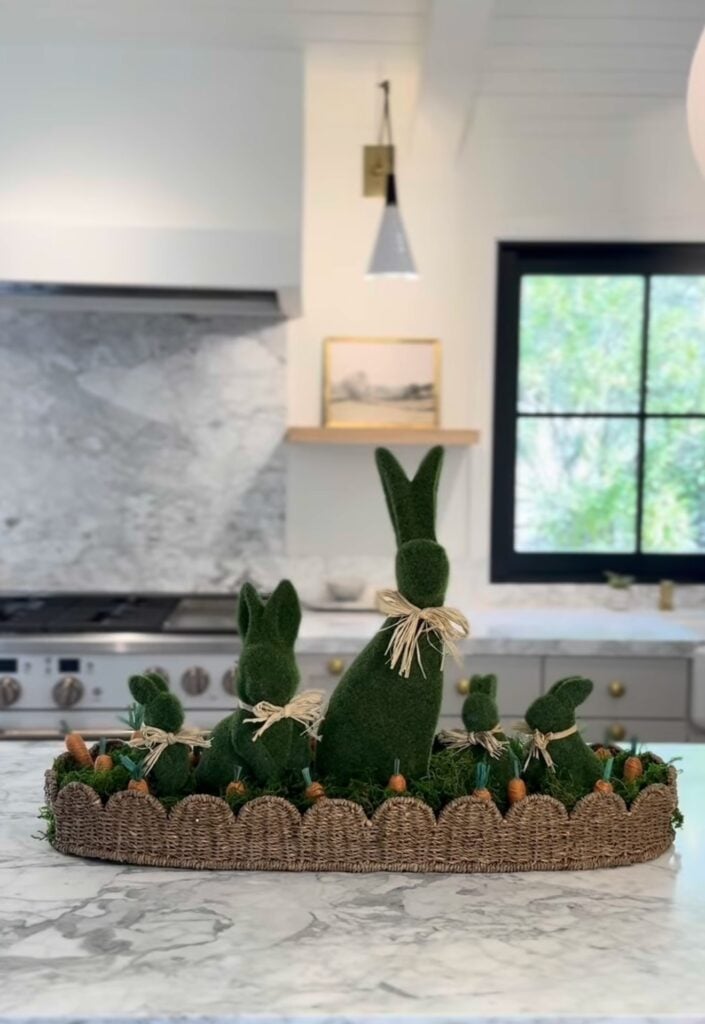 Easter kitchen island centerpiece with green moss bunny topiaries tied with raffia bows in a woven tray accented with moss and tiny carrots.