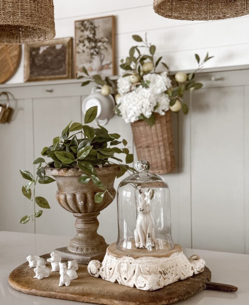 Easter kitchen decor idea with a rustic wood board, leafy planter, white bunny under a glass cloche, and tiny ceramic bunny figurines in a neutral farmhouse kitchen.