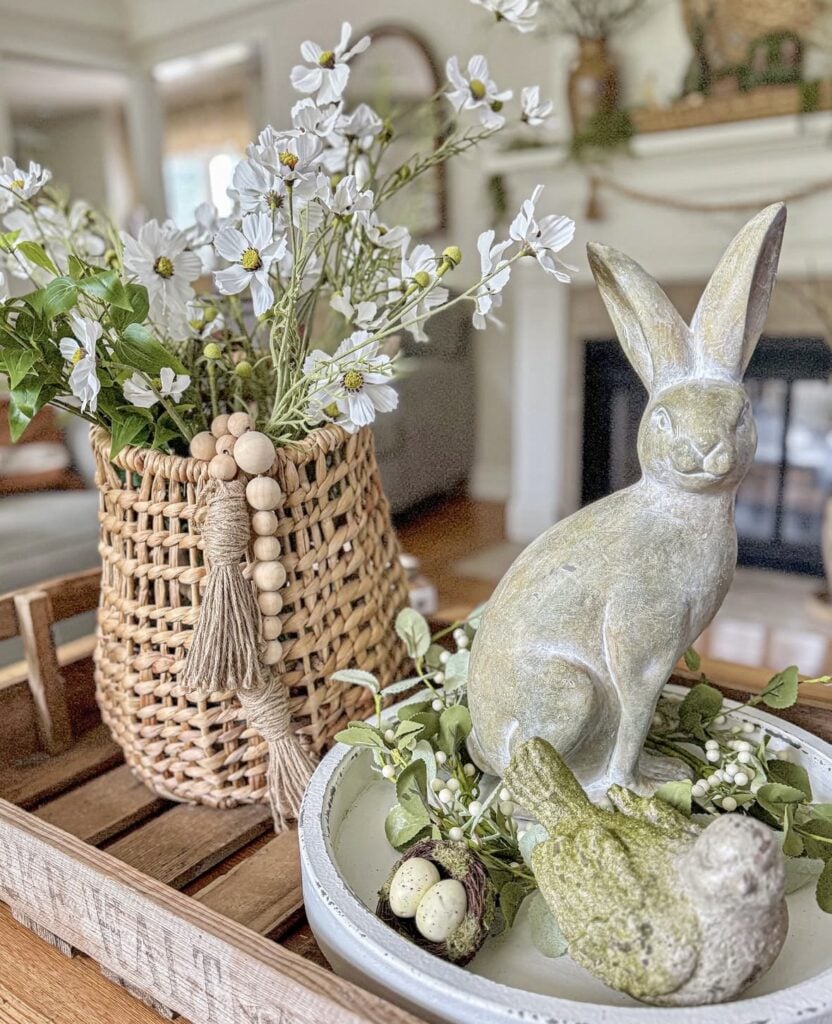 Rustic Easter tray decor with a woven basket of white spring flowers, a stone bunny figurine, a bird statue, and a nest with speckled eggs.