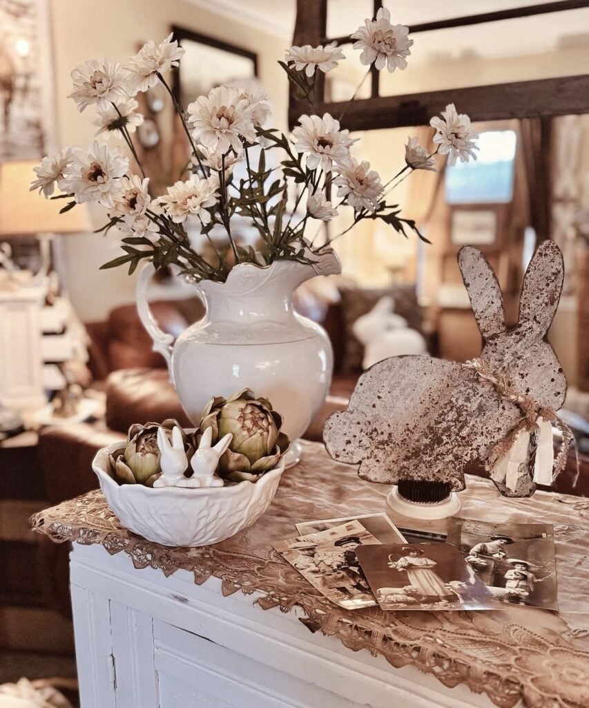 Vintage Easter decor display with a white floral pitcher, distressed bunny figure, ceramic bowl with artichokes and bunny ear accents, and antique photos on a lace runner.
