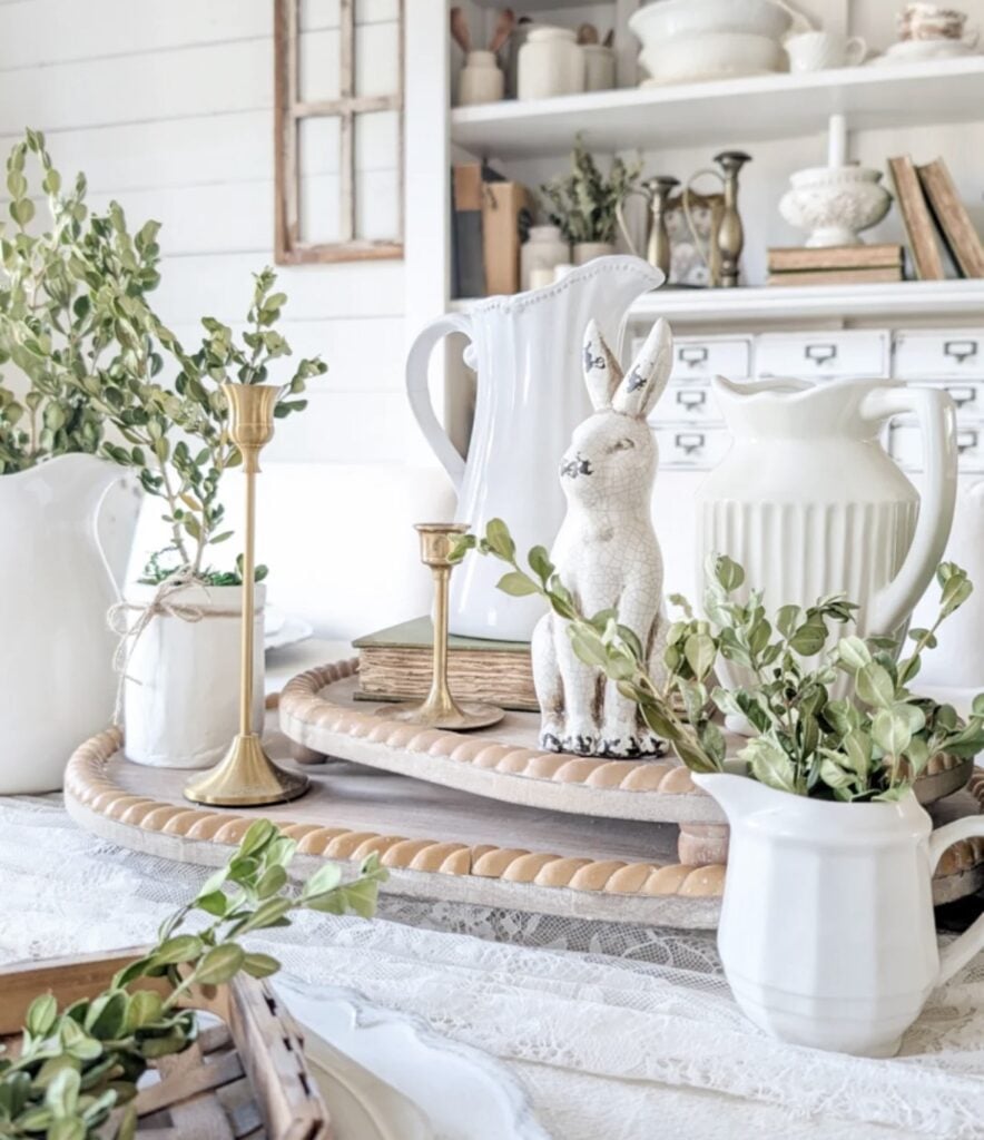Neutral Easter table decor with white pitchers, gold candlesticks, greenery, and a distressed white bunny centerpiece on layered trays.