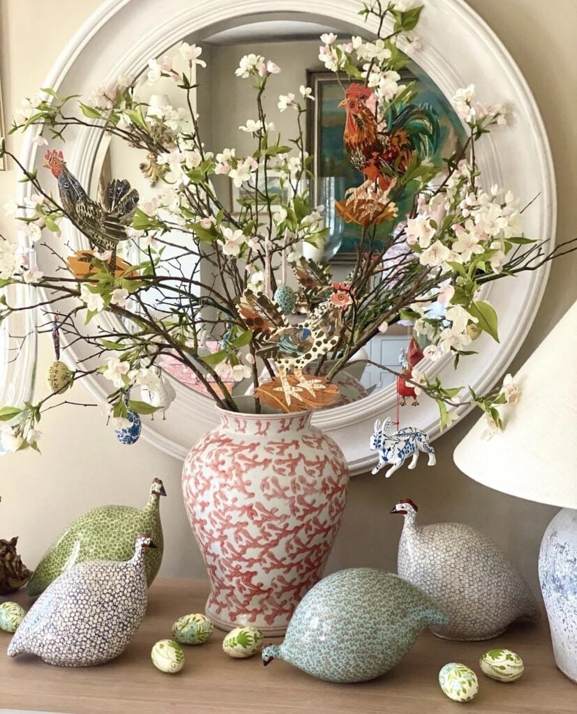 Spring mantel decor with pink blossom branches in a coral pattern vase, colorful rooster accents, hanging Easter ornaments, and decorative guinea fowl figurines.