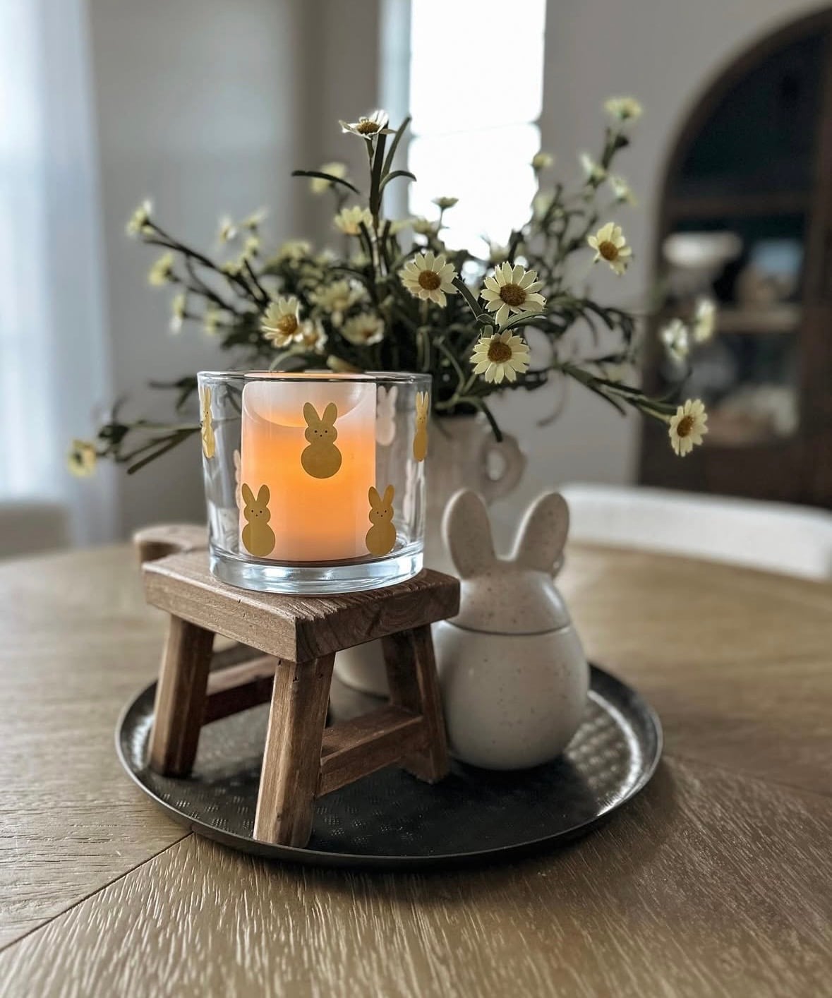 Simple Easter table centerpiece with a bunny Peeps candle holder on a wooden stool, white daisy flowers, and a small ceramic bunny on a black tray.