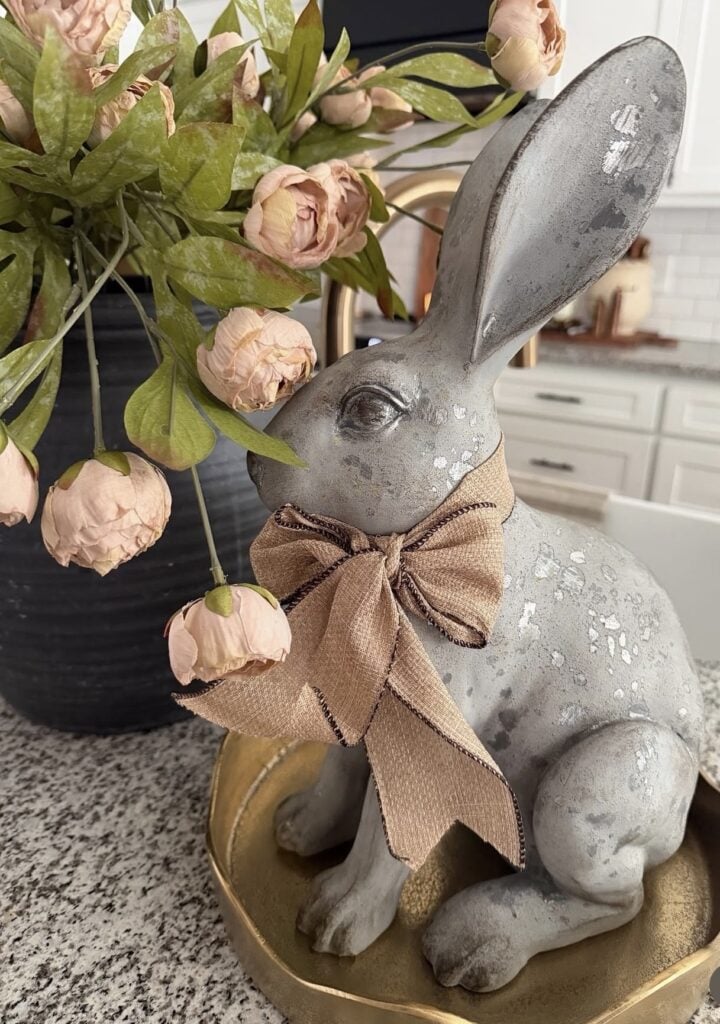 Large distressed gray bunny statue with a tan bow beside a black vase of blush pink flowers on a kitchen counter.