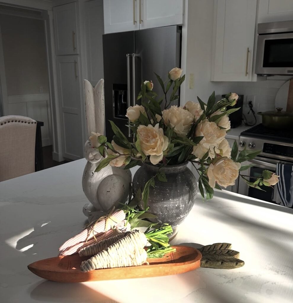 Pink Easter kitchen decor with blush tulips in a glass vase filled with pastel eggs, a gold bunny figurine, and a pink candle in an egg-shaped holder on a round tray.