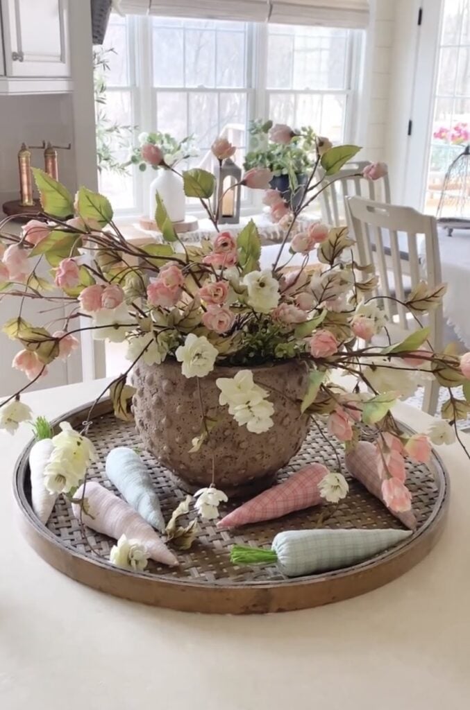 Pastel Easter kitchen island centerpiece with a large floral arrangement in a rustic pot, plaid fabric carrots, and soft spring colors on a round woven tray.
