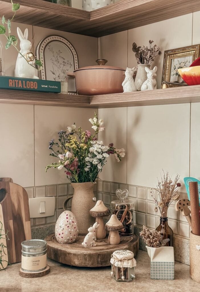 Warm neutral Easter kitchen corner decor with fresh flowers in a vase, bunny figurines, mushroom accents, a floral decorative egg, and wood and glass accessories on a round riser.