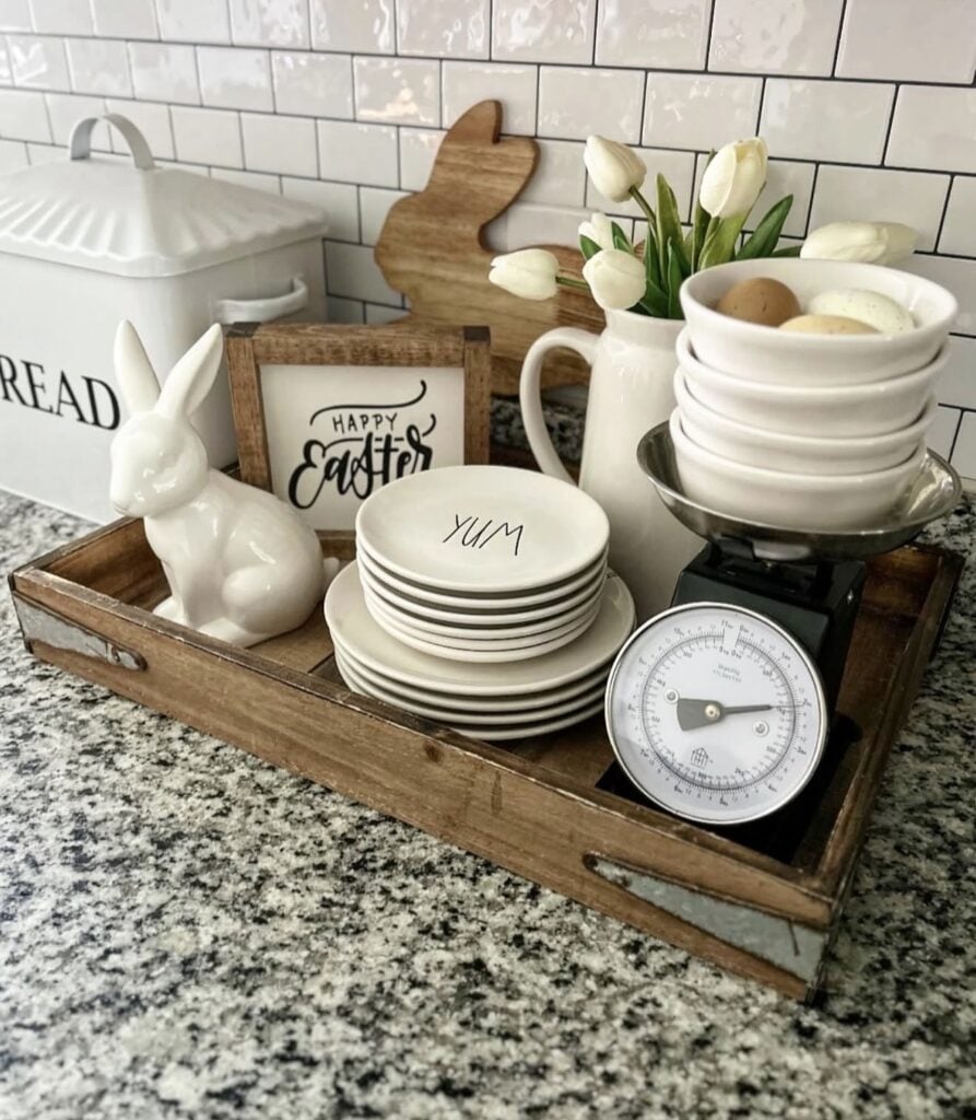 Farmhouse Easter kitchen tray decor with a white ceramic bunny, stacked plates, a Happy Easter sign, white tulips, and bowls of eggs on a vintage kitchen scale.