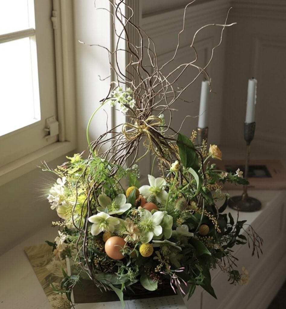 Whimsical Easter floral arrangement with curly branches, white and yellow spring flowers, greenery, and decorative eggs in a rustic nest-inspired centerpiece by a window.