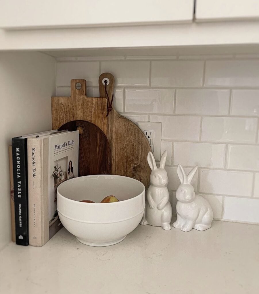 Simple neutral Easter kitchen shelf styling with Magnolia Table cookbooks, layered cutting boards, a white bowl of fruit, and glossy white ceramic bunny figurines.