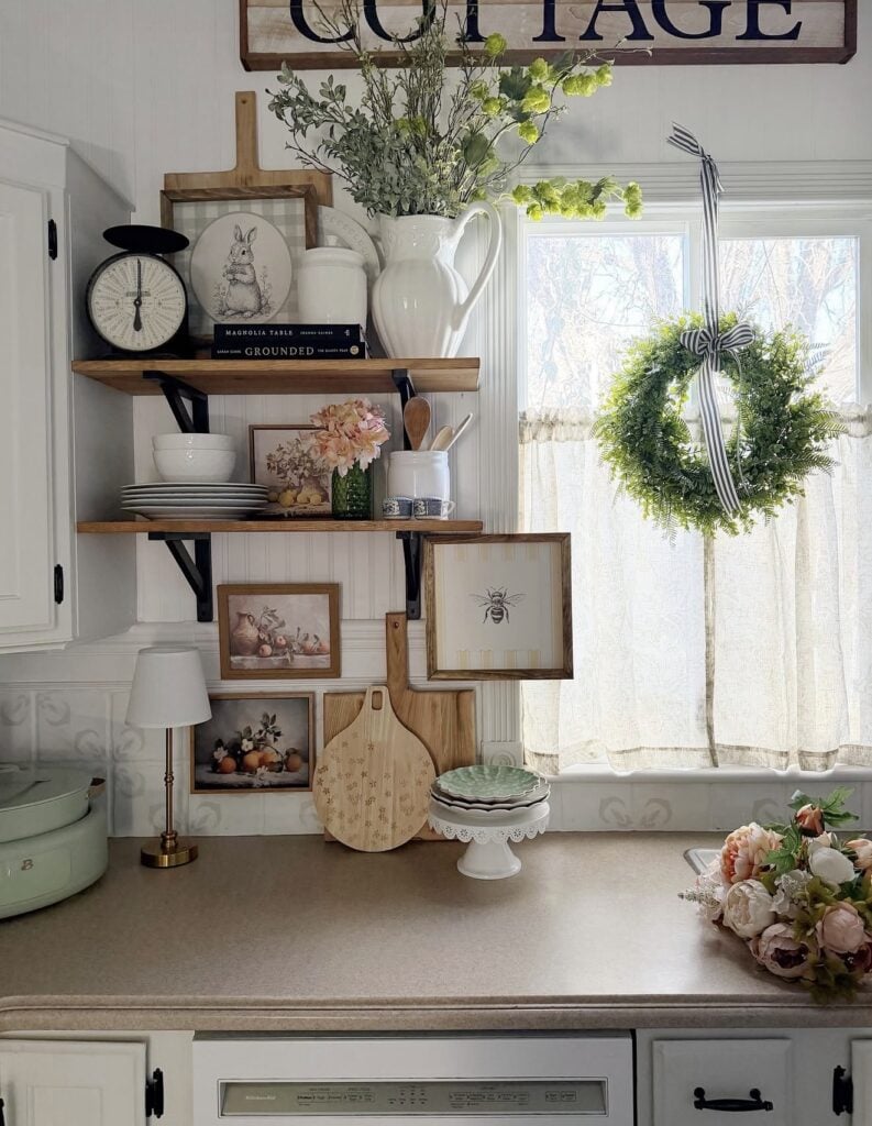 Cottage Easter kitchen decor with open wood shelves, spring florals, framed rabbit and bee art, stacked plates, a green wreath on the window, and soft pastel accents.