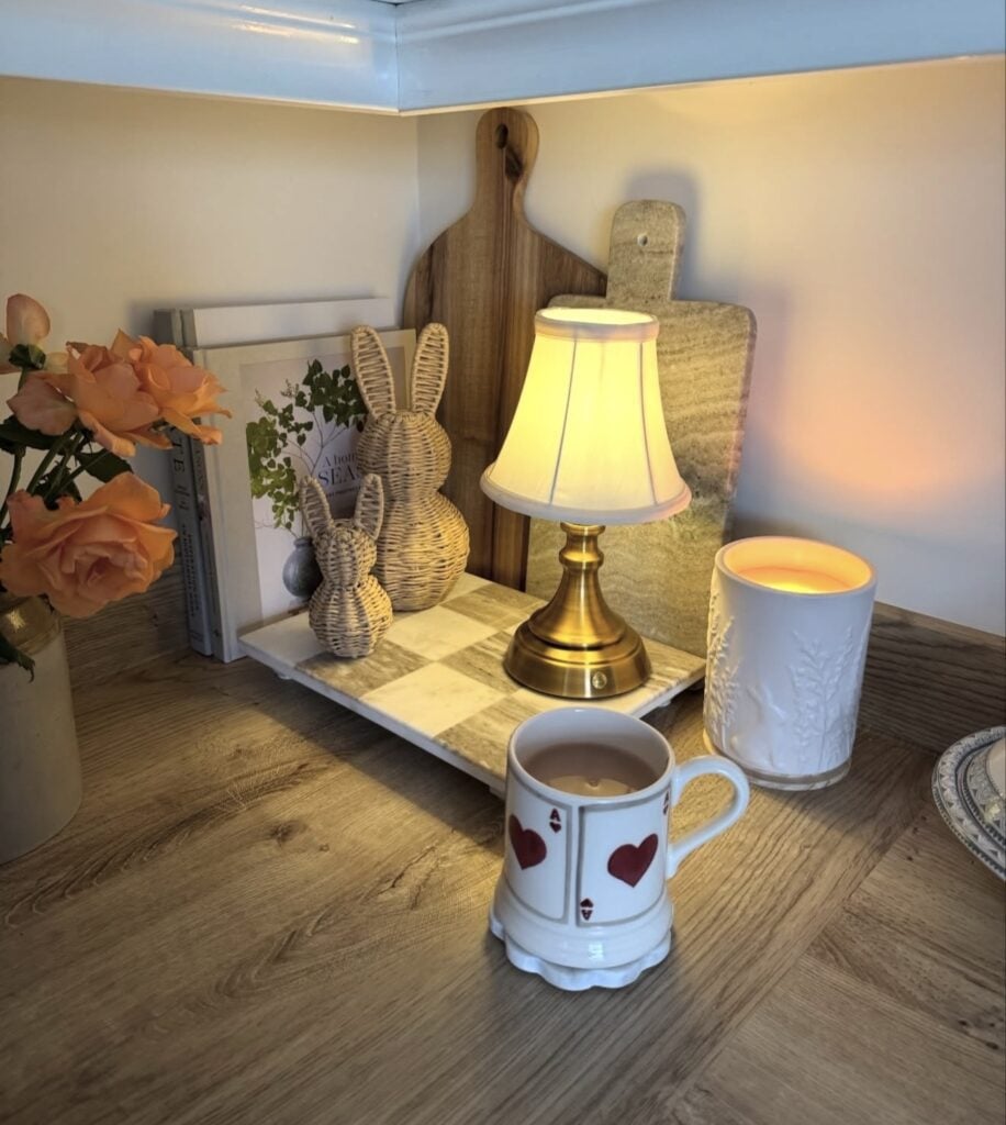 Cozy Easter kitchen nook with wicker bunny figurines, a small gold lamp, a glowing candle, roses, and a heart mug on a marble board.