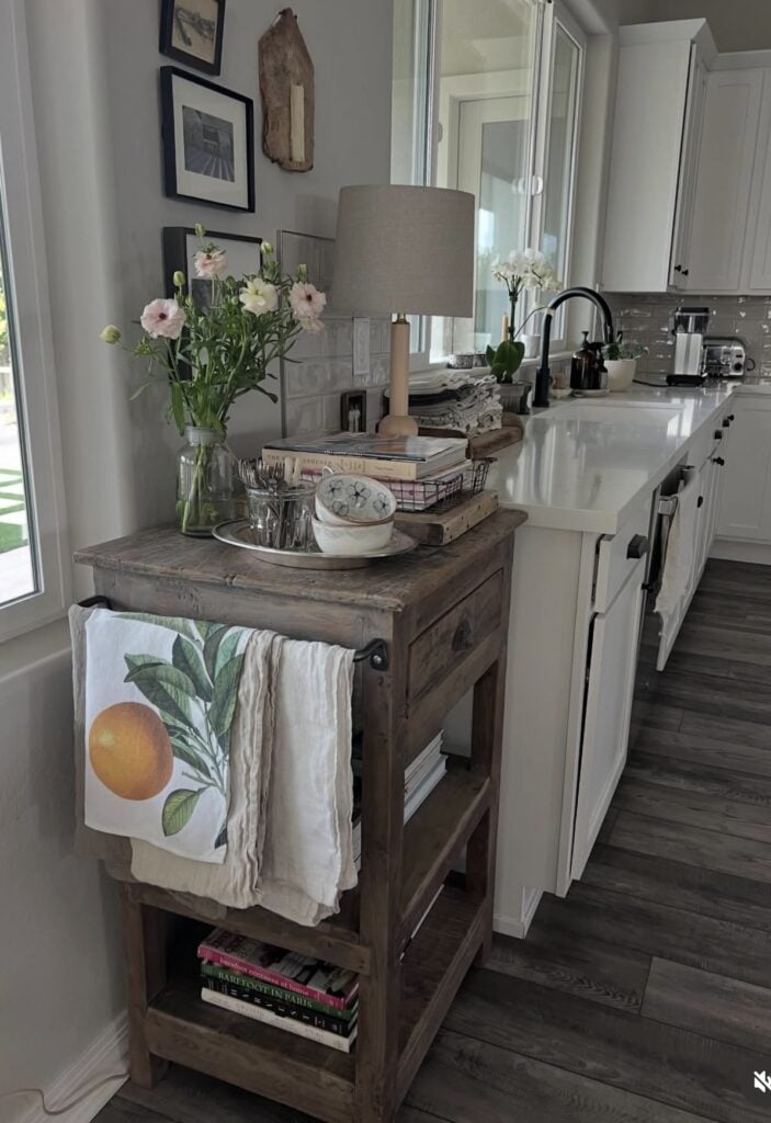 Simple spring kitchen decor on a rustic cart with fresh flowers, stacked books, a small lamp, and citrus-themed dish towels in a bright white kitchen.