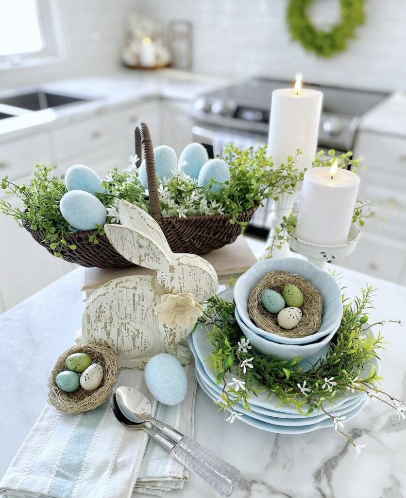 Bright Easter kitchen island decor with a wicker basket of blue eggs, white candles, bird nest bowls, greenery, and a distressed wooden bunny centerpiece.