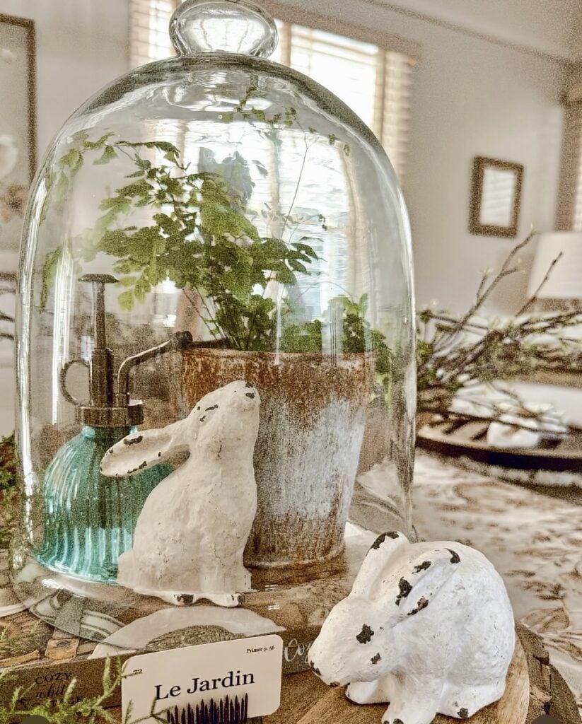 Rustic Easter kitchen decor under a glass cloche with a potted fern, distressed white bunny figurines, and soft vintage spring styling.