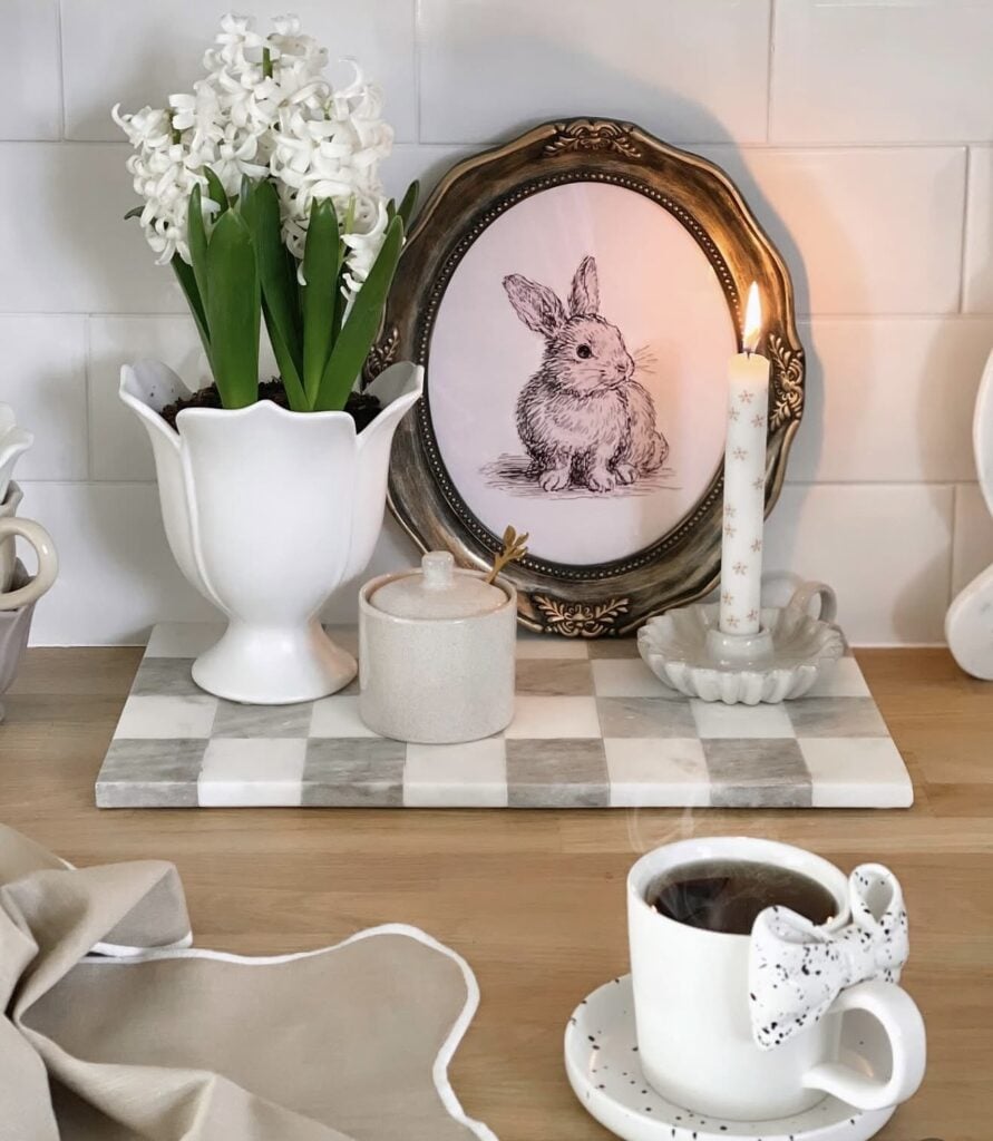 Neutral Easter kitchen decor with white hyacinths in a tulip vase, framed bunny art, a lit taper candle, and a speckled bow coffee mug on the counter.