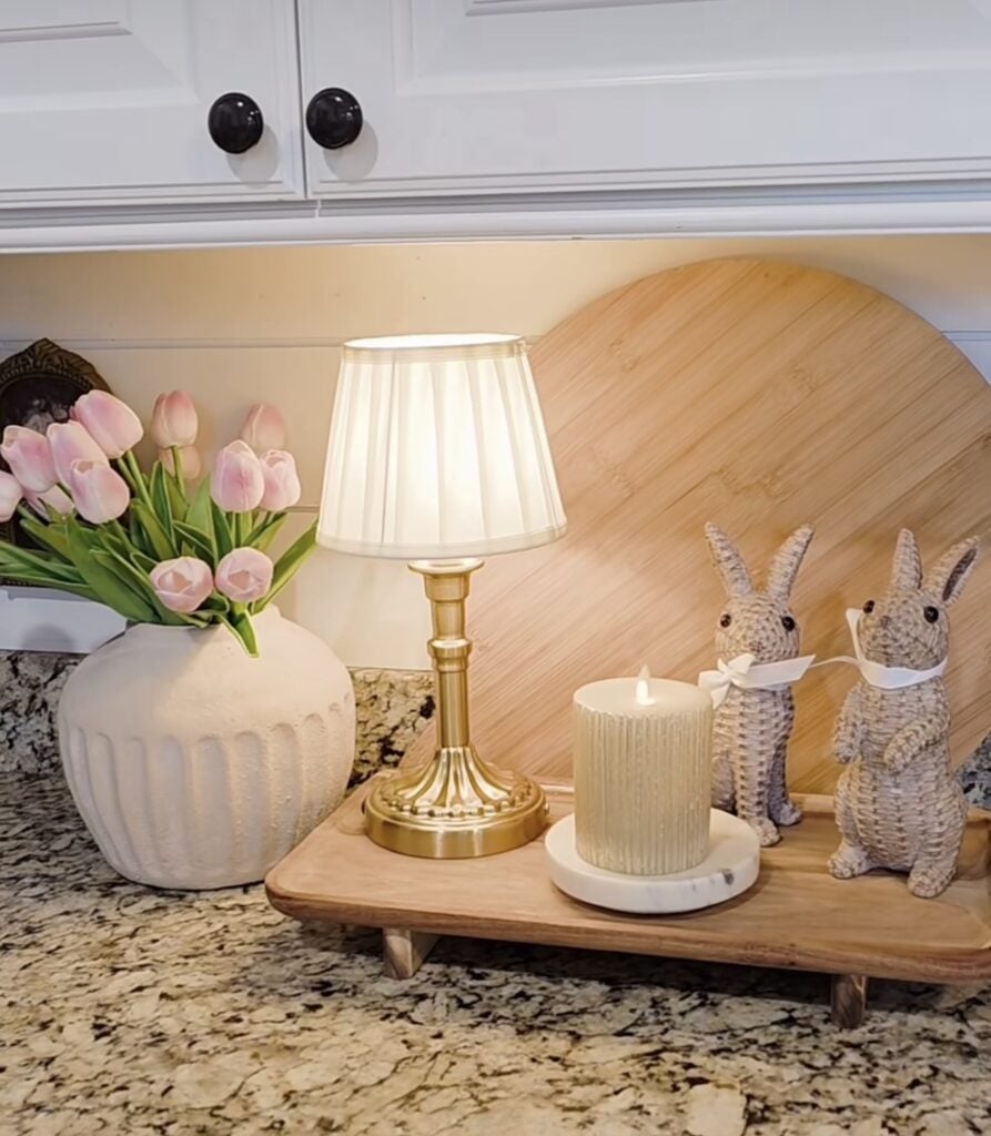 Cozy Easter kitchen counter decor with pink tulips in a textured vase, a small gold lamp, a ribbed candle, and two woven bunny figurines on a wooden riser.