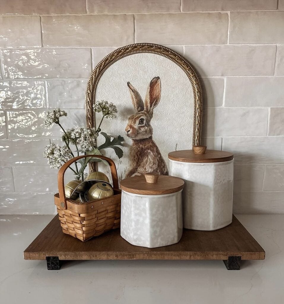 Neutral Easter kitchen decor with a framed rabbit art print, white canisters with wood lids, a small flower vase, and a woven basket on a dark wood riser.