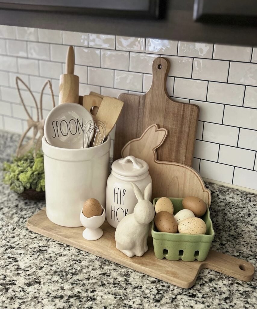 Rae Dunn Easter kitchen decor with a white utensil crock, bunny cutting boards, a Hip Hop canister, decorative eggs, and a small bunny figurine on a wooden board.