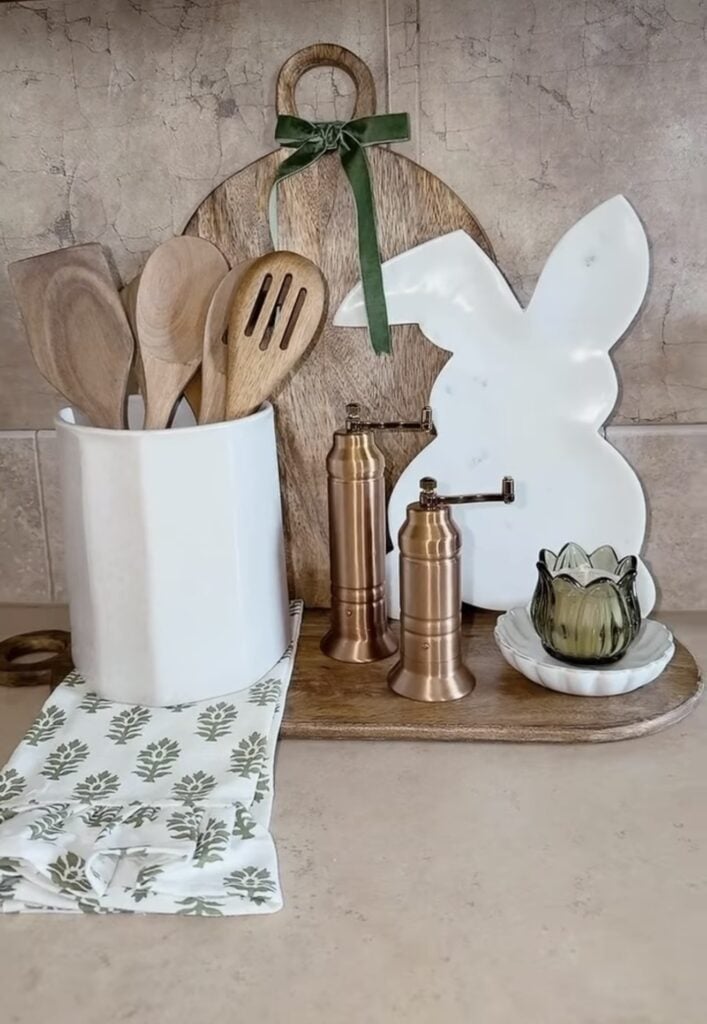 Neutral Easter kitchen decor with a white utensil crock, wooden cutting boards, copper salt and pepper grinders, a white bunny silhouette board, and a green glass candle on a wooden tray.