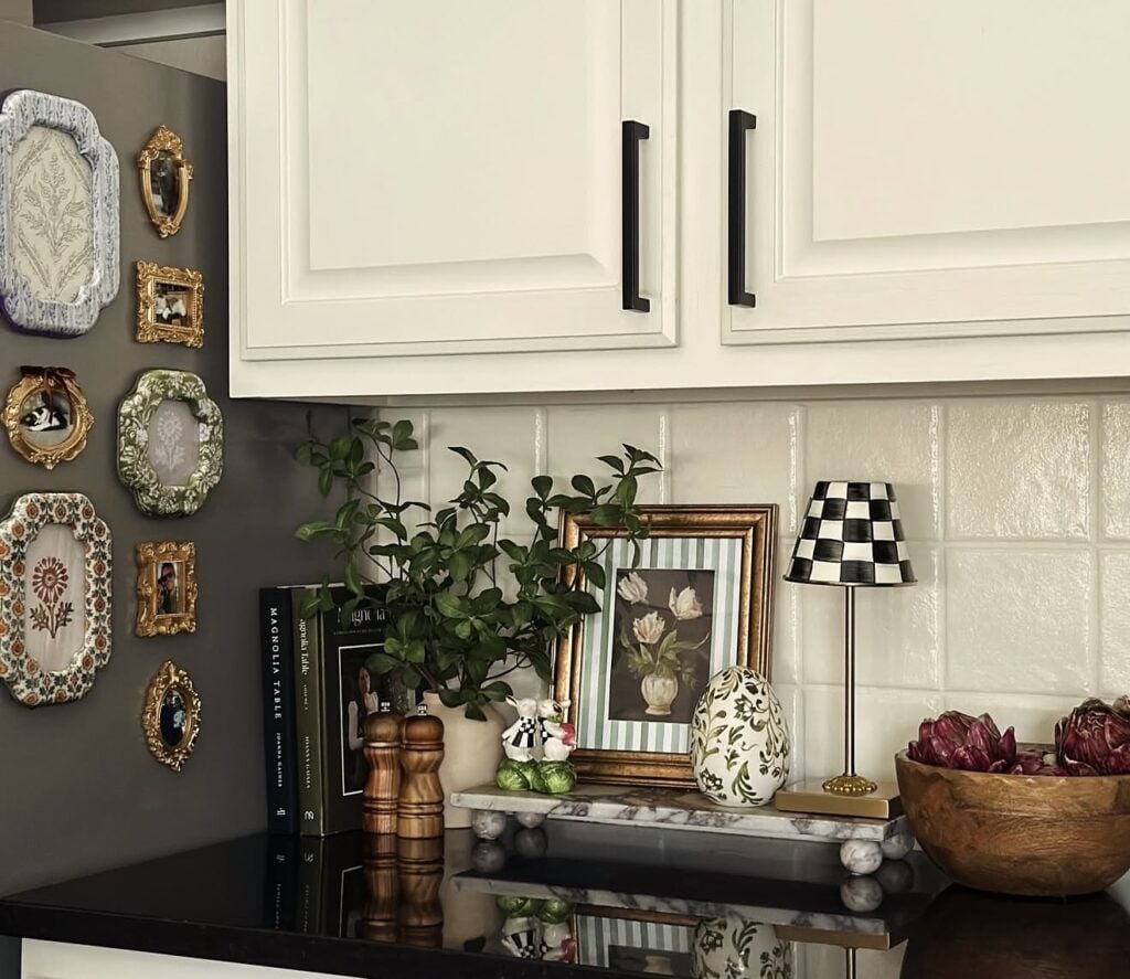 Elegant Easter kitchen vignette with framed art, greenery, books, a floral decorative egg, a small black and white lamp, and a wooden bowl of purple artichokes.