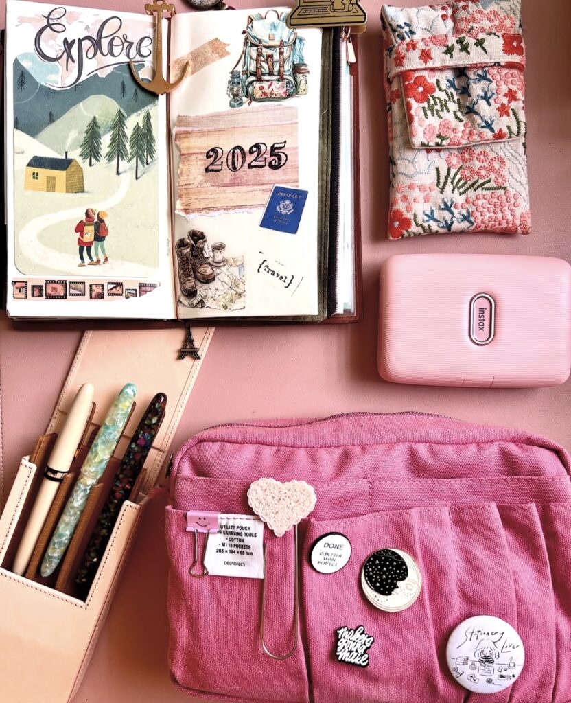Pink desk flat lay with an open travel journal titled “Explore” and “2025,” surrounded by pens, an Instax printer, and floral fabric pouches.
