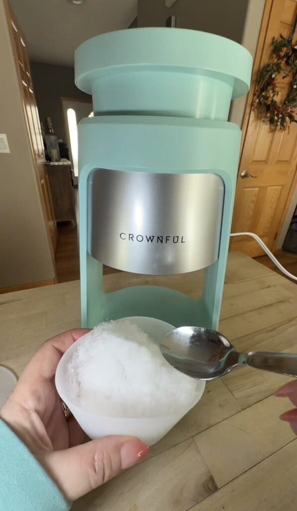 Freshly shaved ice in the small cup in front of the Crownful machine with a spoon ready to use.