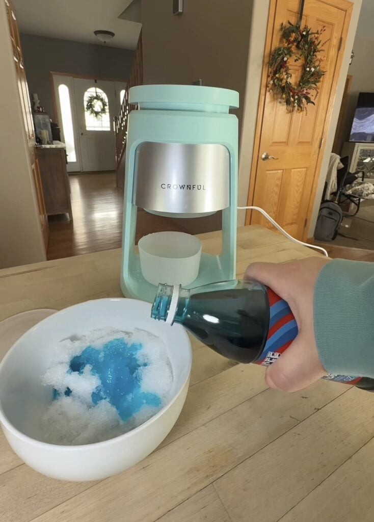 Blue snow cone syrup being poured over shaved ice beside the Crownful shave ice machine.