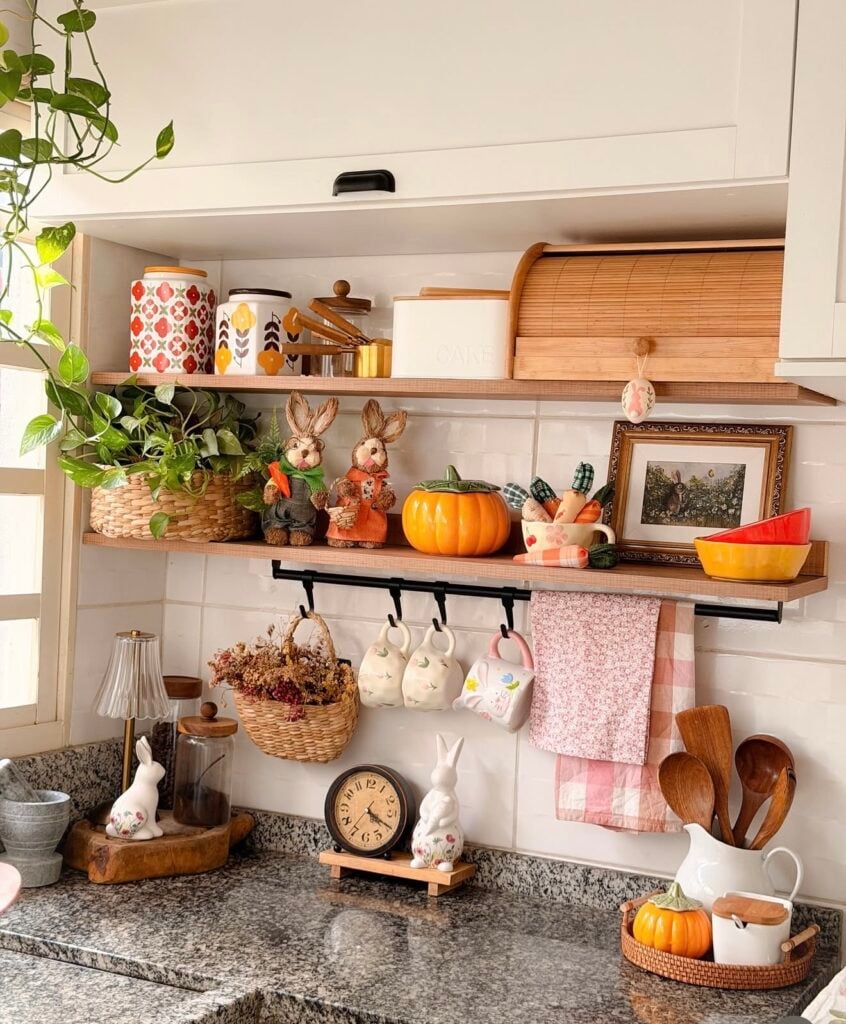 Charming Easter kitchen shelf decor with bunny figurines, pastel mugs, pink dish towels, pumpkins, baskets, and spring accents styled above a granite counter.