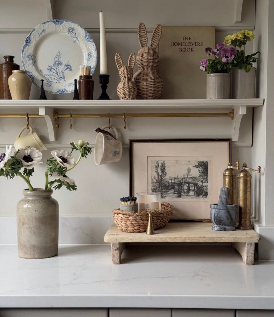 Neutral Easter kitchen counter decor with wicker bunny figurines, a blue and white plate, fresh flowers, vintage art, brass accents, and layered shelf styling.