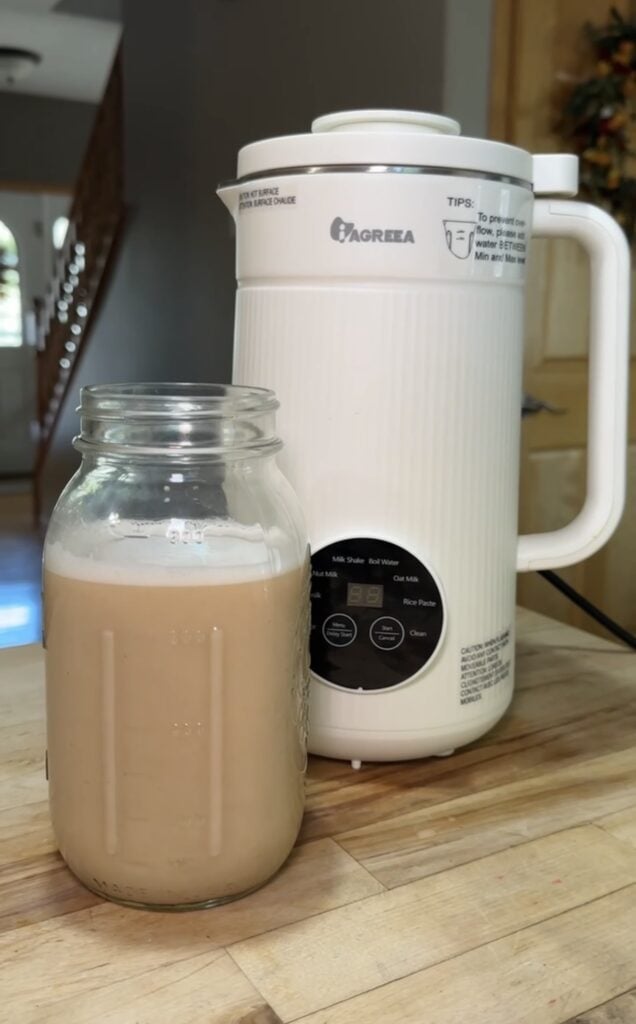 White Iagreea nut milk maker machine on a kitchen counter beside a glass jar filled with freshly made homemade nut milk, featured on slowestuff.com.