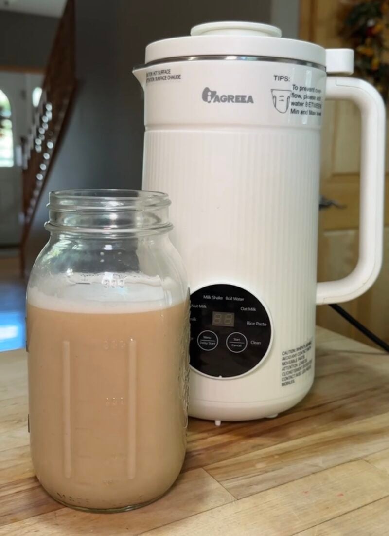 White Iagreea nut milk maker machine on a kitchen counter beside a glass jar filled with freshly made homemade nut milk, featured on slowestuff.com.