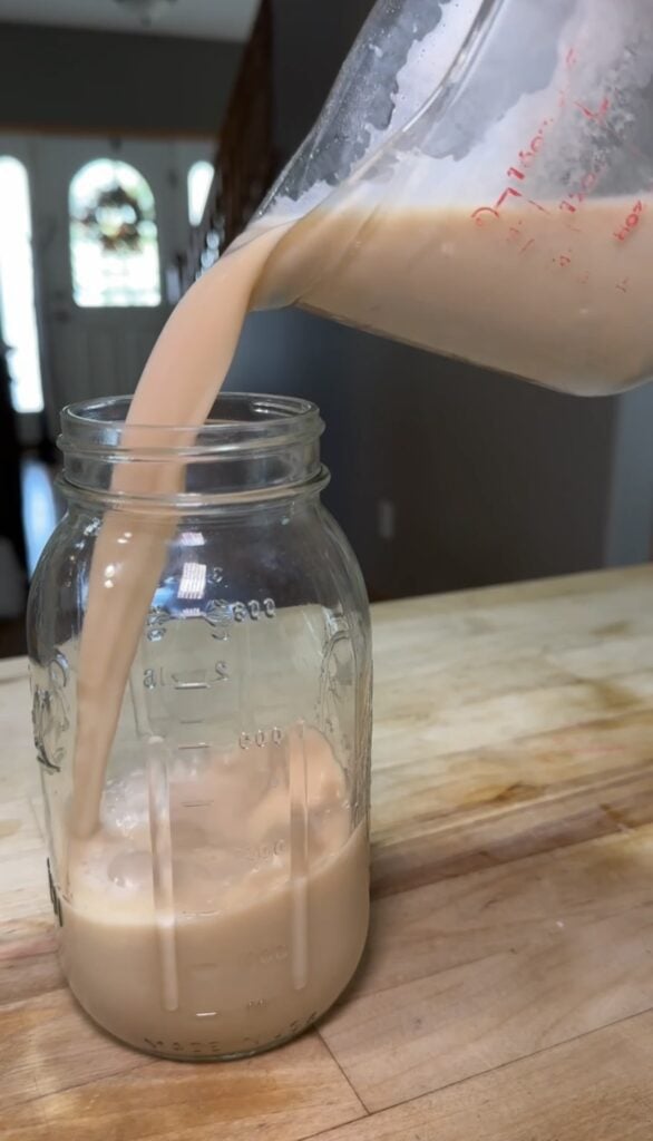 Fresh homemade nut milk being poured from a measuring pitcher into a clear mason jar on a wood kitchen counter, featured on slowestuff.com.